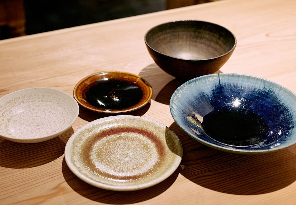 器は5種の釉薬で展開。左上から時計回りに「深鉢中（黒金）」5,500円、「リム皿5寸（飴）」2,750円、「リム皿6寸（胞子）」3,300円、「浅鉢大（天体）」6,600円、「浅鉢小（粉雪）」3,300円 ※（）内は釉薬の種類